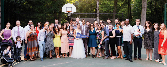 A large line of people, including a bride and groom holding a Brandeis banner, pose outside under a basketball net